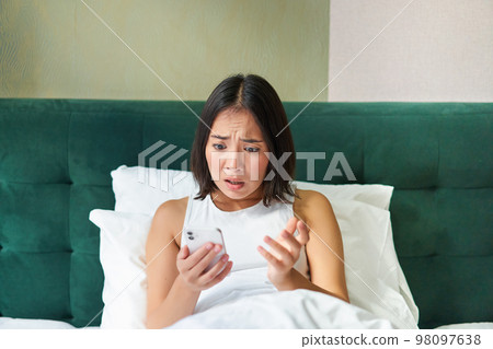 Bedroom picture of asian woman lying in bed, looking scared and shocked at smartphone screen, reading message with concerned face 98097638