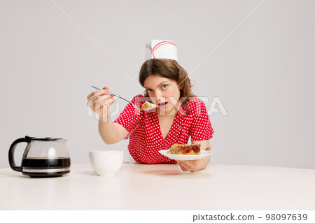 Stylish young female waitress in retro american fashion style of 70s, 80s sitting at table and drinking coffee over grey background. Funny meme emotions, ad, sales Stylish young female waitress in retro american fashion style of 70s, 80s sitting at table and drinking coffee over grey background. Funny meme emotions, ad, sales 98097639
