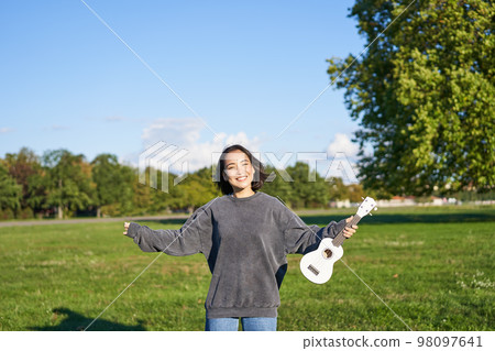 Upbeat young woman dancing with her musical instrument. Girl raises her ukulele up and pose in park on green field 98097641