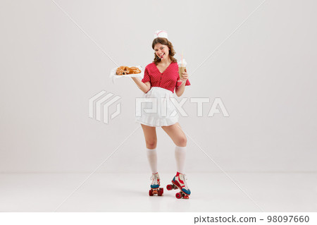 Emotional young woman, retro waitress in american fashion style of 70s, 80s uniform posing over light studio background. Comic photography style Emotional young woman, retro waitress in american fashion style of 70s, 80s uniform posing over light studio background. Comic photography style 98097660