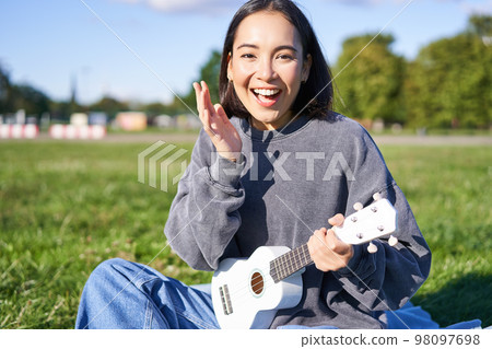Portrait of cute asian girl with musical instrument. Young woman with surprised face, holding ukulele and sitting in park on blanket 98097698