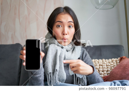Excited young woman pointing finger at smartphone, showing online doctor, medical application or GP contact on mobile phone, staying at home sick, catching cold Excited young woman pointing finger at smartphone, showing online doctor, medical application or GP contact on mobile phone, staying at home sick, catching cold 98097791