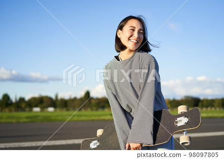 Portrait of asian woman with longboard. Korean girl skating, holding skateboard in hands, posing on road, smiling and looking aside Portrait of asian woman with longboard. Korean girl skating, holding skateboard in hands, posing on road, smiling and looking aside 98097805