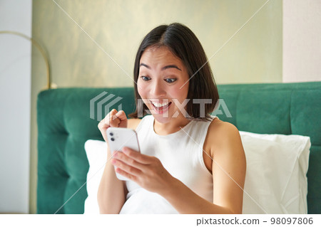 Winning and triumphing. Excited girl in bed, looking at her phone with amazed, surprised face, celebrating victory, reading good news 98097806