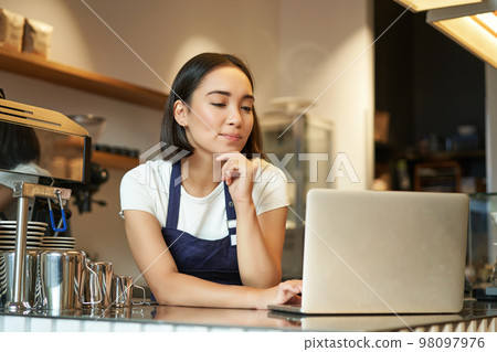 Portrait of smiling korean woman, barista in coffee shop, standing at counter with laptop, smiling and looking confident, self-employed female entrepreneur in her own coffee shop 98097976