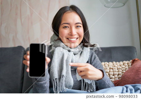 Portrait of smiling girl staying ill at home, showing mobile phone screen, recommending online medical application, pharmacy or gp on smartphone Portrait of smiling girl staying ill at home, showing mobile phone screen, recommending online medical application, pharmacy or gp on smartphone 98098033