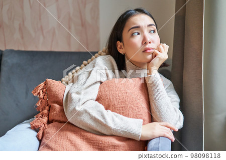 Women and wellbeing concept. Portrait of thoughtful asian woman sitting on sofa with pillow, looking aside with thinking concentrated face 98098118