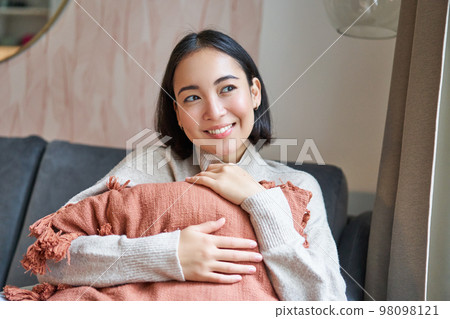 Women and wellbeing concept. Young woman in warm clothes, sits at home, hugs pillow on sofa and smiles pleased, thinks of smth 98098121