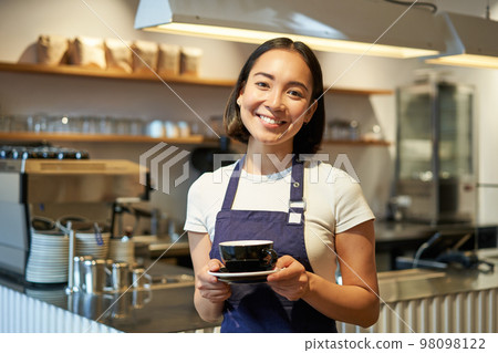 Portrait of smiling asian woman holds cup of coffee, prepare drinks for clients in cafe, working and serving drinks, wearing uniform apron Portrait of smiling asian woman holds cup of coffee, prepare drinks for clients in cafe, working and serving drinks, wearing uniform apron 98098122