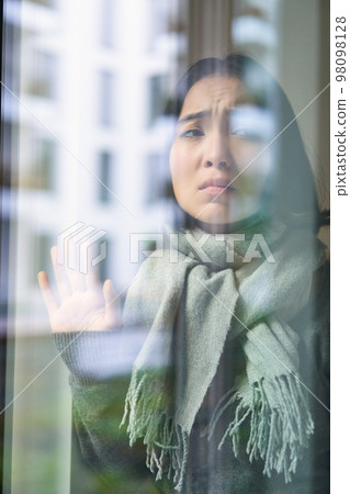 Asian woman feels unwell, stays at home, looks outside window with jealous, distressed face, wants go outside but caught cold 98098128