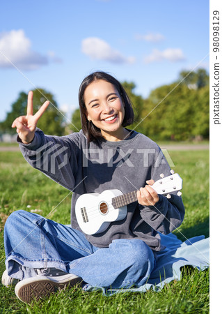 Vertical shot of positive asian girl shows peace sign, plays ukulele in park, rests and enjoys the day. Copy space Vertical shot of positive asian girl shows peace sign, plays ukulele in park, rests and enjoys the day. Copy space 98098129