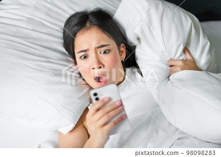 Close up portrait of asian girl lying in bed, looking at smartphone concerned, waking up late and staring at her alarm clock on mobile phone 98098283