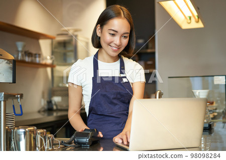 Portrait of young girl cafe owner, looking at her laptop, taking order, serving customer in coffee shop 98098284