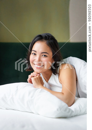 Vertical shot of smiling korean woman lying on pillow in her bed, spending morning in bedroom, waking up from sleep, looking happy 98098503