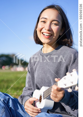Vertical shot of happy korean girl sitting in park, learning how to play ukulele, singing and relaxing 98098586