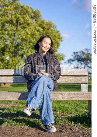 Vertical shot of beautiful brunette woman sitting on bench with smartphone, waiting for someone, using mobile appliation Vertical shot of beautiful brunette woman sitting on bench with smartphone, waiting for someone, using mobile appliation 98098588