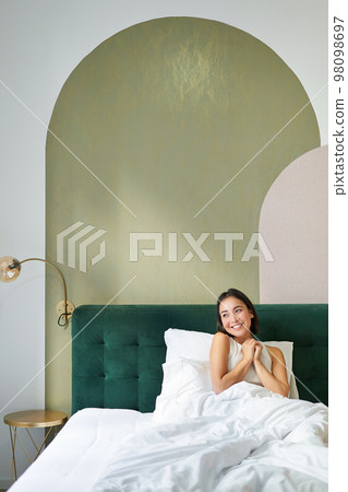 Vertical shot of beautiful asian girl lying in bedroom with sophisticated interrior, smiling and looking happy in morning, spending time in bed 98098697