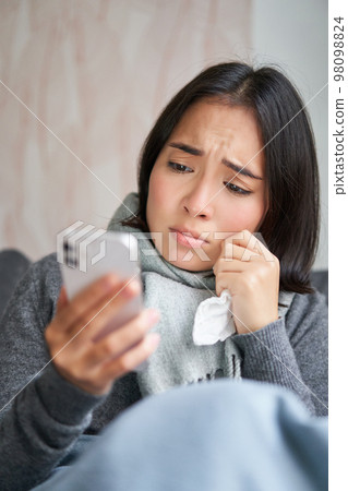 Ill korean woman sitting at home, sneezing, caught cold, reading news on mobile phone, freezing at her home because of heating problems Ill korean woman sitting at home, sneezing, caught cold, reading news on mobile phone, freezing at her home because of heating problems 98098824