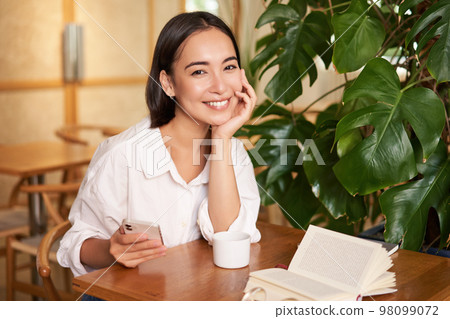 Beautiful young woman with smartphone, drinking coffee in cafe and smiling, relaxing with favorite book 98099072