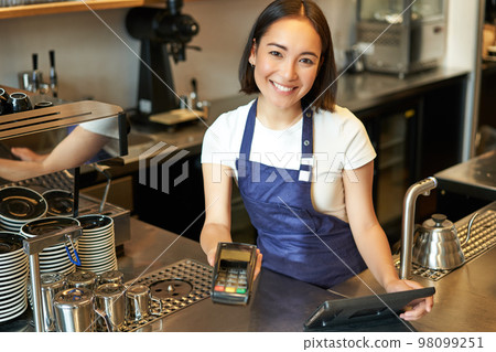Portrait of beautiful asian girl barista, coffee shop staff gives POS terminal, helps pay contactless with credit card, standing at counter in cafe Portrait of beautiful asian girl barista, coffee shop staff gives POS terminal, helps pay contactless with credit card, standing at counter in cafe 98099251
