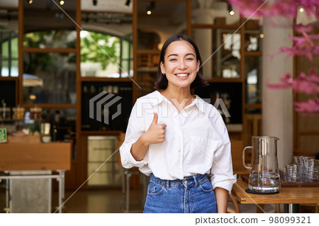 Confident asian businesswoman, showing thumbs up, standing near entrance of her cafe or restaurant, recommending place Confident asian businesswoman, showing thumbs up, standing near entrance of her cafe or restaurant, recommending place 98099321
