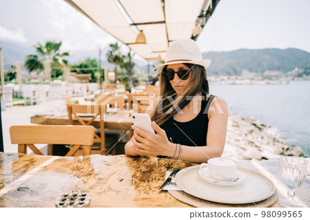 Young girl female lady in sunglasses looking at her mobile smart phone gadget in a street seaside cafe restaurant coffee shop with scenery mountains in the background. Hello summer holiday vacation Young girl female lady in sunglasses looking at her mobile smart phone gadget in a street seaside cafe restaurant coffee shop with scenery mountains in the background. Hello summer holiday vacation 98099565