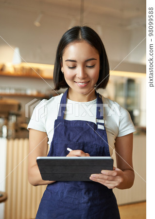 Vertical shot of smiling girl waitress, barista in coffee shop, wears blue apron uniform, takes order with tablet, stands in cafe 98099588