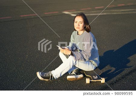 Sad asian teen girl sits on skateboard, looks upset, holds mobile phone and frowns 98099595