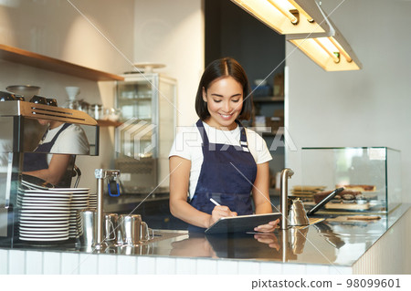 Smiling cute barista girl, korean woman working in cafe, using tablet to process order, making coffee at counter Smiling cute barista girl, korean woman working in cafe, using tablet to process order, making coffee at counter 98099601