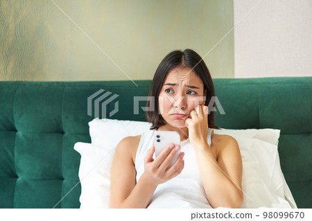 Portrait of sad frowning korean girl, lying in bed and looking at smartphone with upset, disappointed face expression, using mobile phone, complaining 98099670