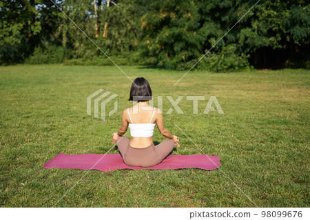 Rear view of woman silhouette doing yoga, sitting on fitness mat and meditating on green lawn 98099676