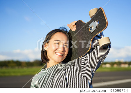 Happy and free asian girl holding cruiser board on shoulders and walking towards camera on empty road, skating on longboard and enjoying sunny weather Happy and free asian girl holding cruiser board on shoulders and walking towards camera on empty road, skating on longboard and enjoying sunny weather 98099724