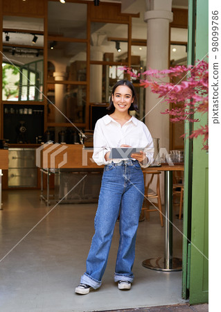 Vertical shot of smiling asian businesswoman, waitress standing in front of cafe entrance, inviting guests in restaurant with tablet Vertical shot of smiling asian businesswoman, waitress standing in front of cafe entrance, inviting guests in restaurant with tablet 98099786