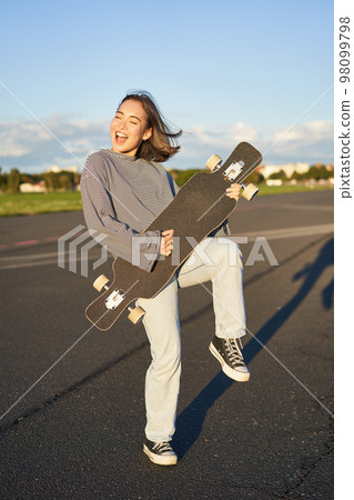 Funny asian girl enjoying skating, holding skateboard like guitar and shadow playing, having fun outdoors 98099798