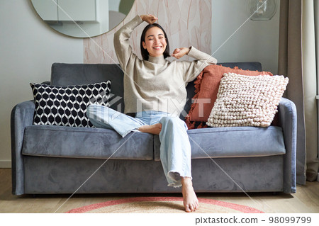 Portrait of happy asian woman feeling lazy, stretching on sofa and smiling pleased, relaxing at home, resting from work 98099799