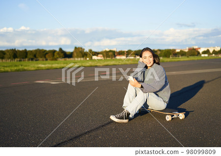 Portrait of young korean girl sitting on her skateboard on road, looking at smartphone, chatting on mobile app Portrait of young korean girl sitting on her skateboard on road, looking at smartphone, chatting on mobile app 98099809