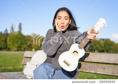 Portrait of young girl musician, sitting in park with ukulele guitar, looking surprised at camera, saying wow 98099844