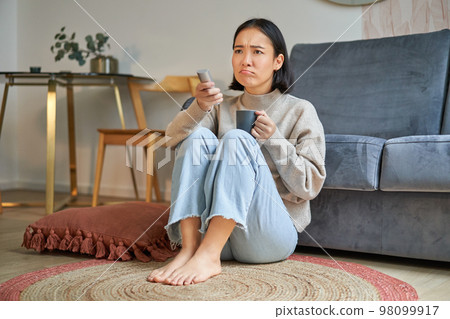 Portrait of young woman with remote, watching tv, switching chanels on television, sitting on floor near sofa and relaxing Portrait of young woman with remote, watching tv, switching chanels on television, sitting on floor near sofa and relaxing 98099917