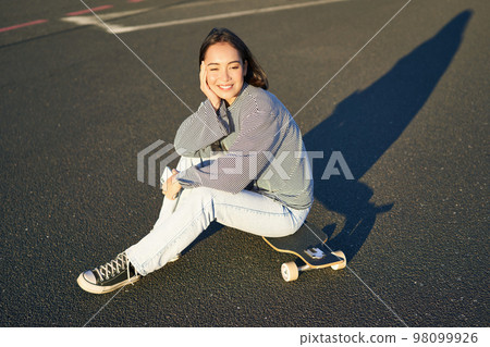 Portrait of asian woman sitting on skateboard, skating on her cruiser longboard, using smartphone app Portrait of asian woman sitting on skateboard, skating on her cruiser longboard, using smartphone app 98099926