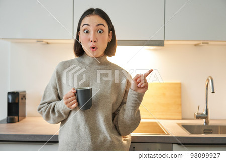 Portrait of smiling asian girl standing in kitchen, drinking coffee from cup and pointing at banner, showing letting agencies advertisement Portrait of smiling asian girl standing in kitchen, drinking coffee from cup and pointing at banner, showing letting agencies advertisement 98099927