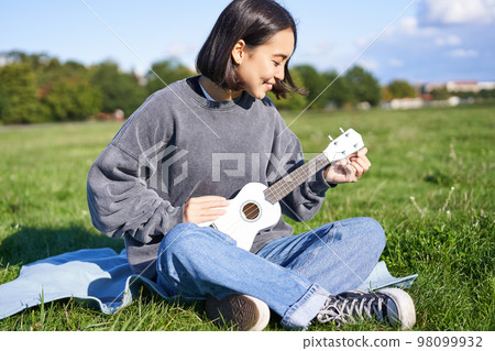 Asian smiling girl tuning her ukulele guitar, singing and playing in park, sitting on blanket on sunny day Asian smiling girl tuning her ukulele guitar, singing and playing in park, sitting on blanket on sunny day 98099932