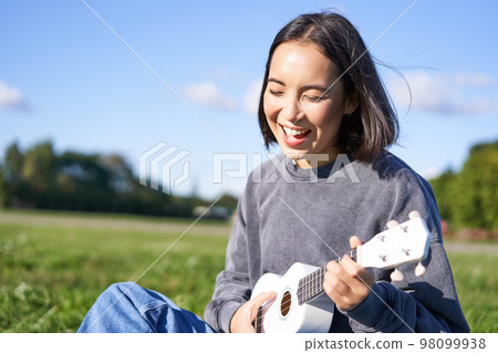Happy people and hobbies. Smiling asian girl playing ukulele guitar and singing, sitting in park outdoors on blanket 98099938