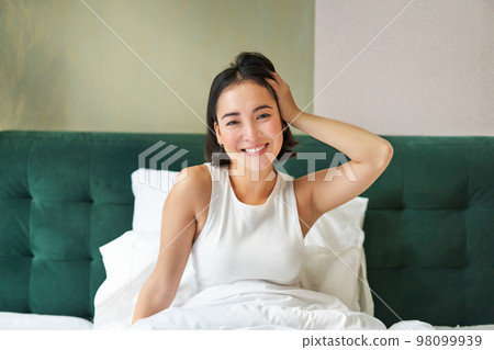 Cute korean girl in white tank top, wakes up in her bedroom, lying in bed in morning, stretching hands and smiles 98099939
