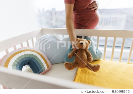 Pregnancy. Pregnant woman preparing nursery holding belly baby bump by crib holding teddy bear. regnancy concept and home nusery planning 98099945