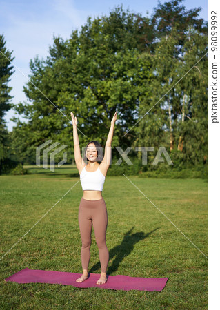 Vertical shot of asian woman raising hands up to sky, meditating, practice yoga, doing wellbeing training in park on green lawn 98099992