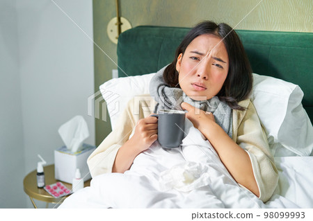 Portrait of couching asian girl in bed, feeling sick, catching cold and staying at home, looking unhealthy, drinking hot tea 98099993