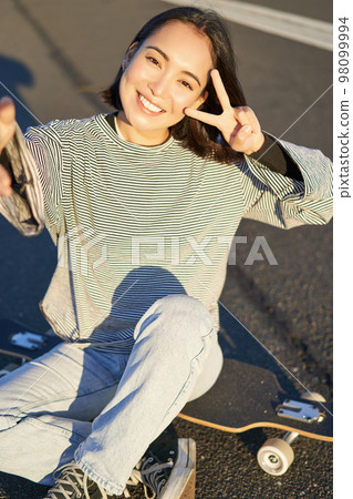 Selfie of asian girl sitting on skateboard, taking photo on smartphone, smiling and showing peace v-sign 98099994
