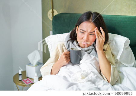 Flu, sickness and people. Young woman feeling sick, having fever and catching col, lying in bed with hot tea, taking drugs from influenza 98099995