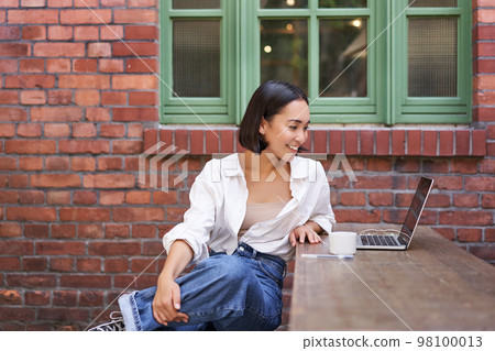 Portrait of young stylish woman, influencer sitting in cafe with cup of coffee and laptop, smiling and looking confident 98100013