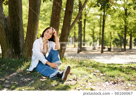 Young people. Beautiful asian girl sits near tree in park and rests, smiling and looking into distance, relaxing outdoors on fresh air in summer Young people. Beautiful asian girl sits near tree in park and rests, smiling and looking into distance, relaxing outdoors on fresh air in summer 98100067